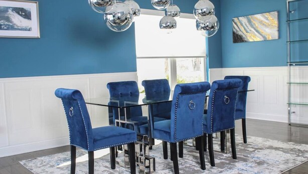 Een eethoek met een glazen tafel, omringd door zes beklede blauwe stoelen, een kamerbreed tapijt en een plafond met reflecterende, bolvormige lampen.