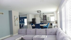 Een moderne woonkamer met een hoekbank, een eethoek met blauwe stoelen, en een keuken met witte kastjes en een betegelde achterwand.