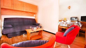 Una sala de estar con un sofá oscuro, una mesa de madera con dos vasos y una tetera, una silla roja y una kitchenette con un microondas y un televisor.