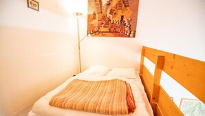 A small, single-bed room with a wooden headboard, white bedding, and a patterned throw blanket. A wall-mounted lamp and a framed picture are visible. - 100m from ski slopes, 2nd floor, balcony, parking, tv, 47m², La Norma (La Norma)