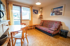A cozy living room with a wooden dining table, chairs, a patterned sofa, and a small shelf with decorative items.