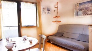 Una habitación acogedora con un sofá gris, una mesa de comedor de madera con sillas, un estante pequeño en la pared y una ventana con cortinas.