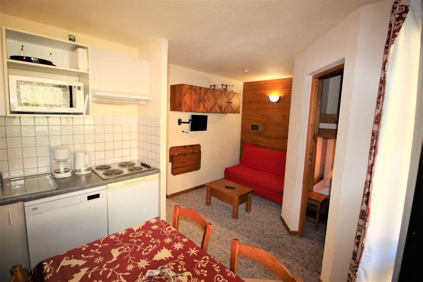 Eine kleine Küche mit Mikrowelle, Kaffeemaschine und Herd. Ein Esstisch mit rotem Tischläufer und Stühlen steht bereit. Eine rote Couch lehnt an der Wand, darüber befindet sich ein Holzschrank.