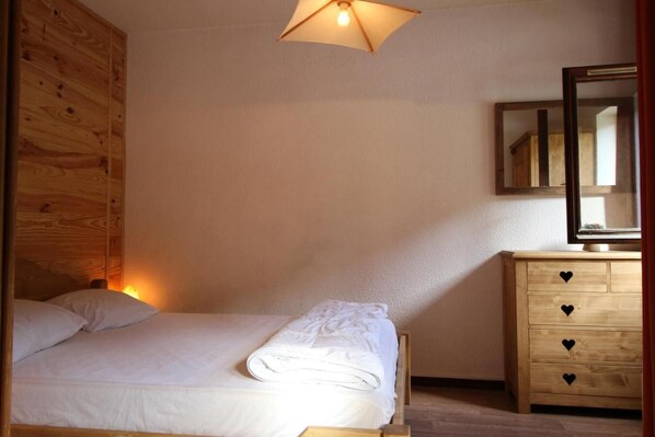 Une chambre à coucher avec une tête de lit en bois, un lit recouvert de draps blancs, une commode en bois dotée de poignées en forme de cœur et un miroir placé au-dessus de la commode.