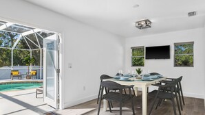 A dining area with a table set for four, a pool visible through an open door, and a television mounted on the wall. - Sunny House with heated pool, 12min drive to Engelwood Beach, Port Charlotte FL (Port Charlotte)