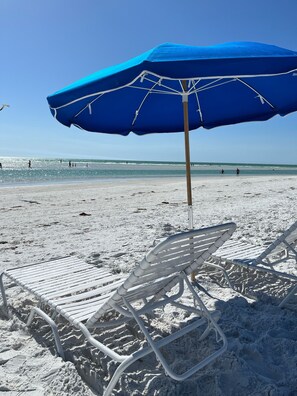 A beach scene with a blue umbrella and white lounge chairs.
