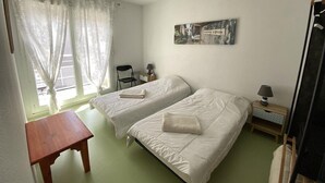A room with two beds, a small table, a chair, a lamp, and a picture on the wall.