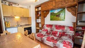 Ein gemütlicher Raum mit einem Holztisch, einer Couch mit Patchworkdecke sowie einer Küchenzeile mit Holzausstattung und einem Gemälde einer Berglandschaft.