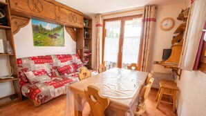Ein gemütliches Zimmer mit einer Holzsofa, einem Esstisch mit Stühlen, einem Holzkasten mit einem Gemälde und einem Fenster mit Vorhängen.