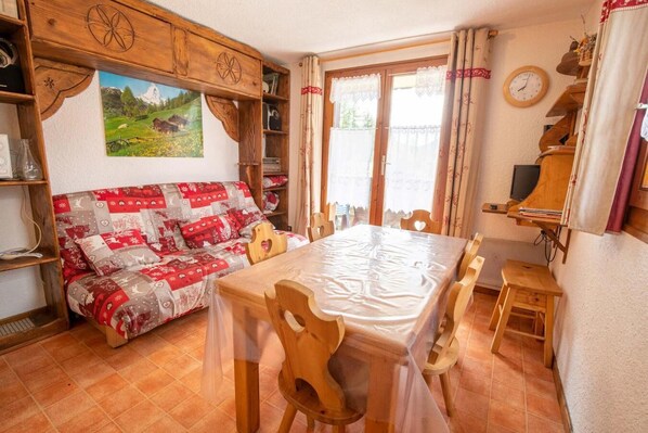 Una habitación acogedora con un sofá de madera, una mesa de comedor con sillas, un mueble de madera con un cuadro y una ventana con cortinas.