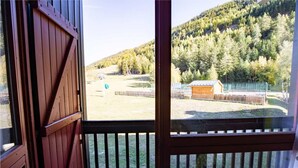 Vista desde un balcón de madera con baranda metálica, que da a un espacio al aire libre cercado con un campo cubierto de césped y un edificio en la distancia.