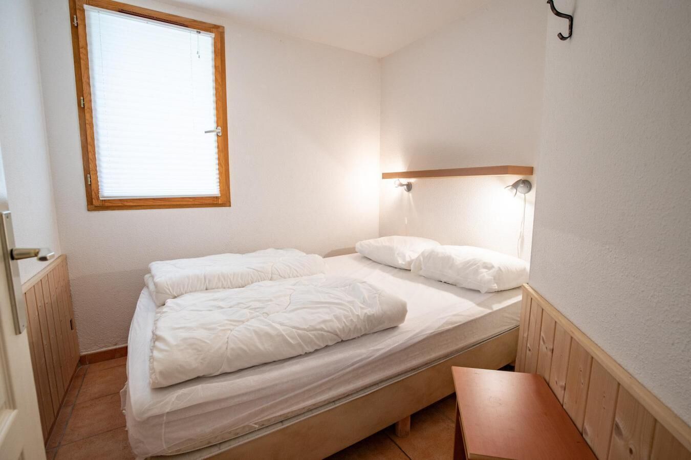 A small, single-bed room with a wooden headboard, a bedside table, and a window with blinds.