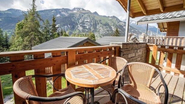 A wooden deck with wicker chairs and a table, overlooking a mountainous landscape.