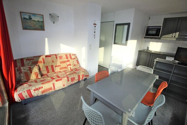 Un espacio habitable compacto con sofá, mesa de comedor, sillas, kitchenette y un ventanal pequeño.
