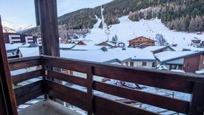 A wooden balcony overlooking a snow-covered village with traditional buildings and a ski slope in the background.