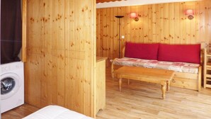 A room with wooden walls, a bed, a sofa with red cushions, a wooden bench, and a washing machine. - Ski-in/ski-out & foot of the slopes, 1st floor, swimming pool, sauna, balcony, tv, ski locker, 35m² (Les Orres)