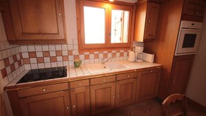 Een keuken met houten kastjes, een betegelde spatwand, een raam met een lamp, een fornuis, een spoelbak en een koffiezetapparaat.