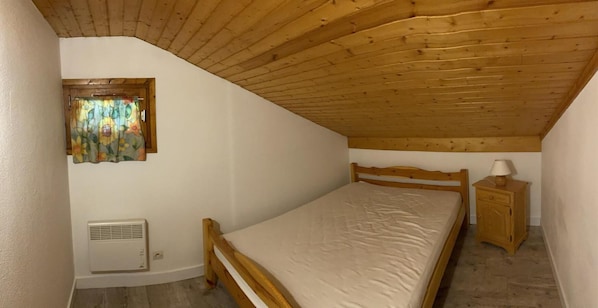 Une petite chambre à coucher accueillante, avec un plafond en bois, un lit simple, une table de chevet et une fenêtre ornée d’un rideau à motifs floraux.