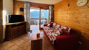 A cozy living room with a wooden floor, a red sofa, a television on a wooden cabinet, and a balcony with a view of mountains. - 200m from shops, 1st floor, swimming pool, balcony, tv, 46m², Les Saisies (Villard-sur-Doron)