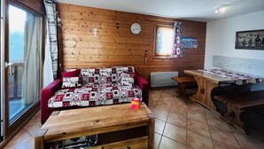 A cozy room with a wooden wall, a red sofa, a wooden table, and a window with curtains. - 200m from shops, 1st floor, swimming pool, balcony, tv, 46m², Les Saisies (Villard-sur-Doron)