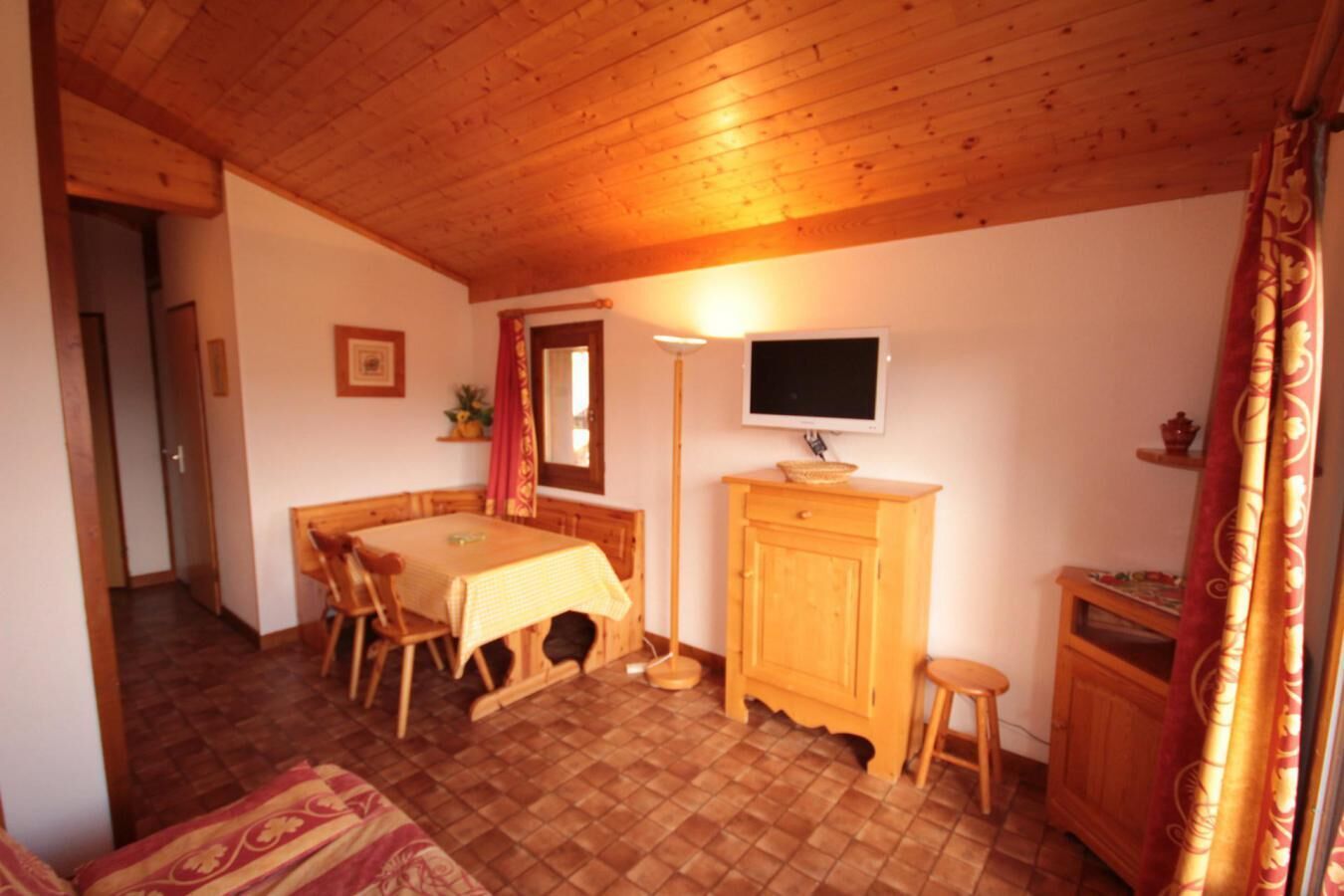 A cozy room with a wooden ceiling, a dining table with chairs, a wooden cabinet, a television, and a patterned curtain.