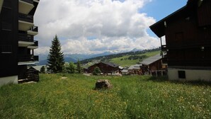 A rural landscape with a modern building on the left, traditional wooden structures in the middle, and a clear view of distant mountains under a partly cloudy sky. - 300m from ski slopes, Ground floor, view mountain, balcony, tv, 28m², Les Saisies (Les Saisies)