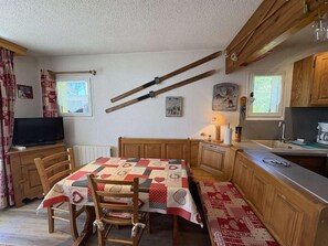 A cozy kitchen with a wooden table, chairs, and a bench. There is a television on a cabinet, and a window with curtains.