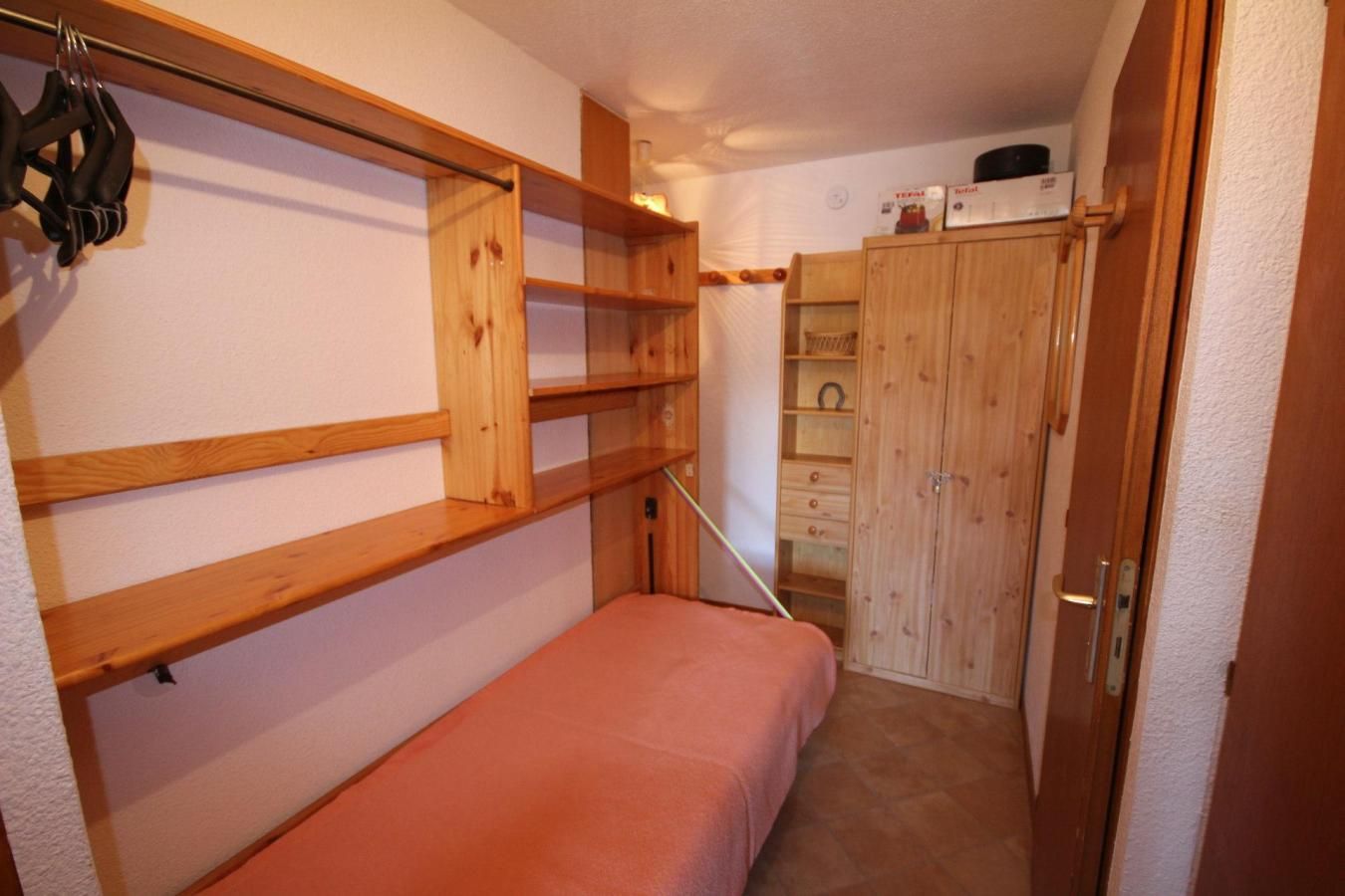A compact room with a single bed, wooden shelves, a wardrobe, and a small table.