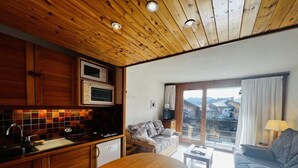 A modern kitchen with wooden cabinets, a microwave, and a dining area with a wooden table. There is a sofa and a window with a view of buildings. - 100m from ski slopes, 1st floor, view ski slopes, balcony, tv, ski locker, 23m², Les Saisies (Les Saisies)