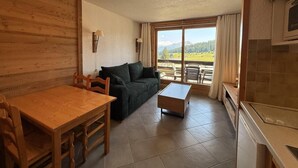 A cozy living room with a wooden dining table, a sofa, and a coffee table. There is a kitchen area with a microwave and cabinets. A large window offers a view of the outdoors. - 200m from ski slopes, 1st floor, view ski slopes, balcony, tv, ski locker, 24m², Les Saisies (Les Saisies)