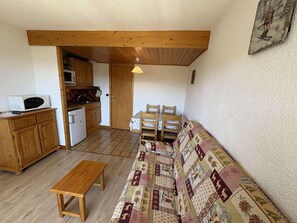 A compact living space with a wooden bench, a patterned sofa, a dining table with chairs, and a kitchen area with a microwave and dishwasher.
