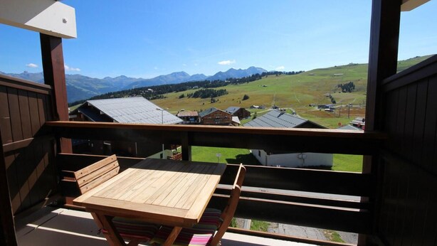Un balcon avec une table et des chaises en bois, donnant sur un paysage montagneux parsemé de bâtiments.