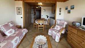 Ein gemütliches Wohnzimmer mit einer Couch, einem Sessel und einem kleinen Tisch mit einer Dekoration. Ein Holzkabinett und ein Fernseher auf einem Schrank sind ebenfalls vorhanden.