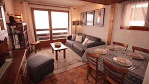 A cozy living room with a sofa, a dining table set for four, and a large window with curtains. - 100m from ski slopes, 1st floor, view ski slopes, balcony, tv, ski locker, 26m², Les Saisies (Les Saisies)