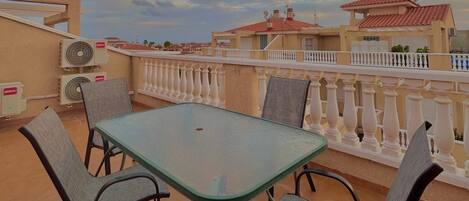 A balcony with a glass table and chairs, air conditioning units, and a white balustrade.