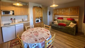 A compact kitchen with wooden cabinets, a microwave, and a small refrigerator. There is a dining table with a checkered tablecloth and chairs. A green sofa with red cushions is in the living area. - 250m from ski slopes, 1st floor, view mountain, balcony, tv, 28m², Les Saisies (Les Saisies)