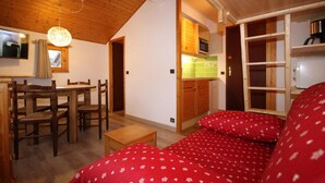 Ein gemütliches Zimmer mit Holzbalkendecke, einem Esstisch mit Stühlen, einer kleinen Küchenzeile mit einer Mikrowelle und einem Bett mit roter Bettwäsche.