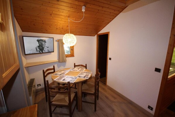 A dining area with a round wooden table, four chairs, a hanging light fixture, a framed picture on the wall, and a door leading to another room.
