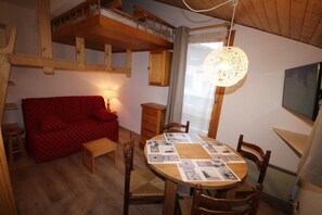 A cozy room with a wooden ceiling, a red sofa, a wooden dining table with a placemat, and a hanging light fixture.