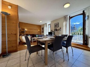 A dining area with a wooden table and chairs, a kitchen in the background, and a view of mountains through a large window.