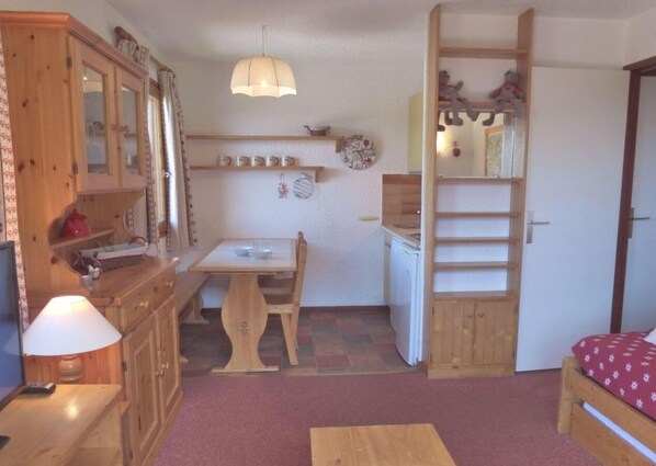 Una habitación compacta con una mesa de comedor de madera, un pequeño área de cocina, una cama con colcha estampada roja y un mueble de madera con puertas de cristal.