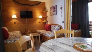 Une pièce chaleureuse avec un mur en bois, une télévision, deux tables de chevet avec des lampes, un lit avec une literie à motifs, une table ronde en bois et une chaise.