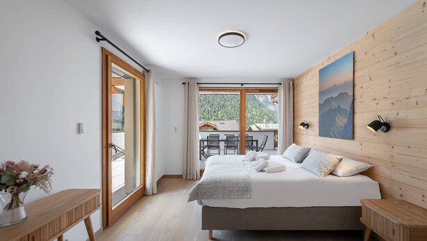 Ett sovrum med en stor säng, träpanel på väggarna, utsikt över bergen och en balkong med utemöbler.