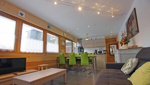 A modern living room with a sofa, a TV, and a dining area with green chairs.