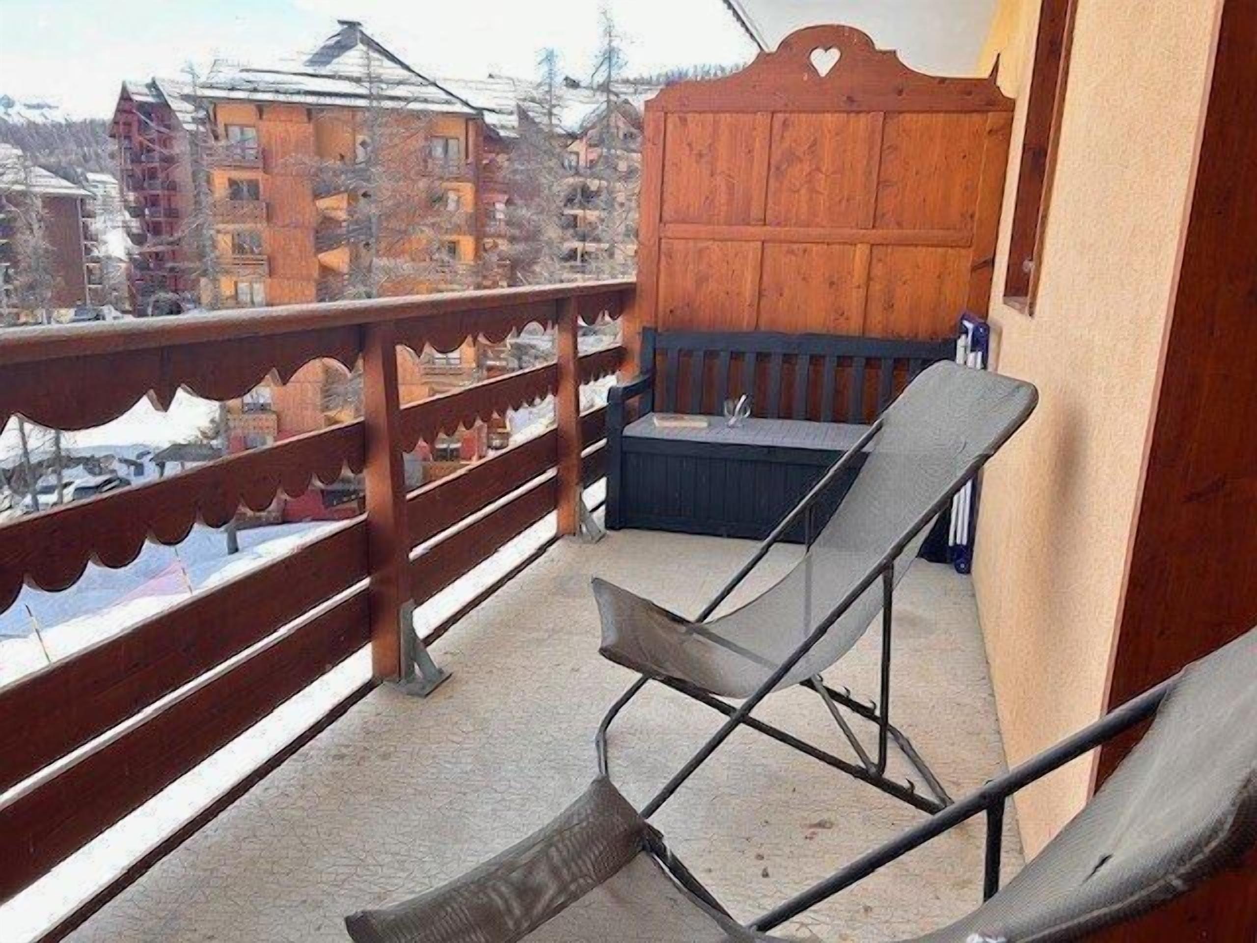 Un balcón con barandilla de madera, una silla negra y una pequeña mesa con una botella.