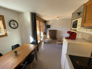 A compact kitchen with a microwave, a red coffee maker, and a small refrigerator. There is a dining area with a wooden table and chairs, and a living space with a sofa and a television.
