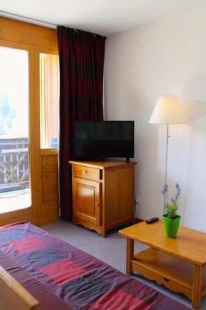 Une pièce avec un lit, un téléviseur posé sur une commode en bois, une lampe et une plante.