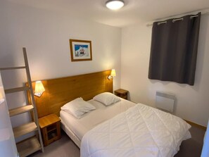 A hotel room with a wooden headboard, a bed with white bedding, a nightstand with a lamp, a small wooden stool, a framed picture on the wall, and a window with a dark curtain. - 50m from ski slopes, 4th floor, view mountain, tv, 40m², Superdévoluy (Le Dévoluy)