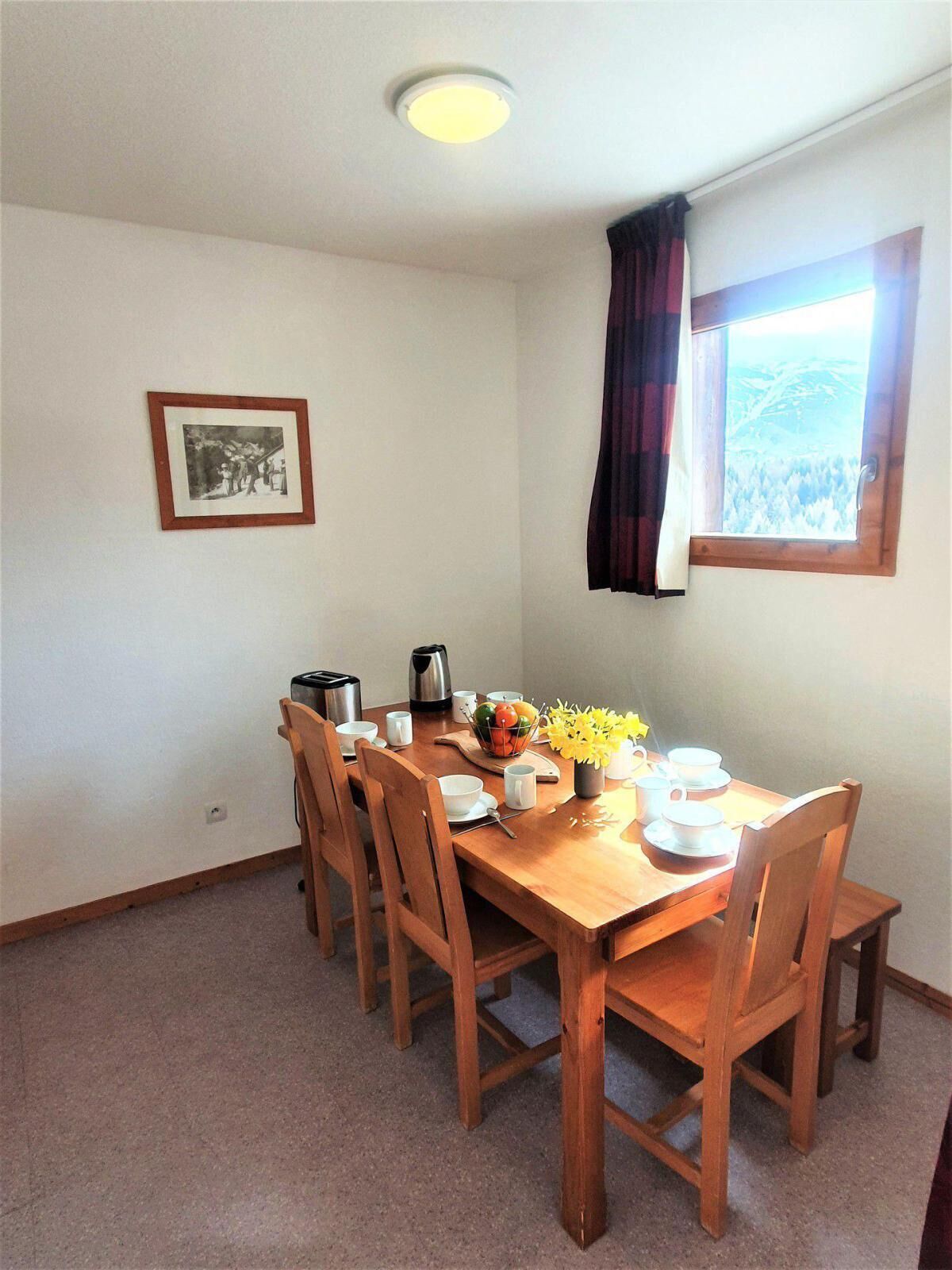 A dining area with a wooden table set for four, surrounded by chairs. There is a framed picture on the wall, a window with curtains, and a view of a mountainous landscape.
