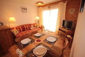 Un accogliente soggiorno con un tavolo da pranzo in legno apparecchiato per quattro persone, un divano con una coperta a quadri, una televisione e un orologio appeso al muro.